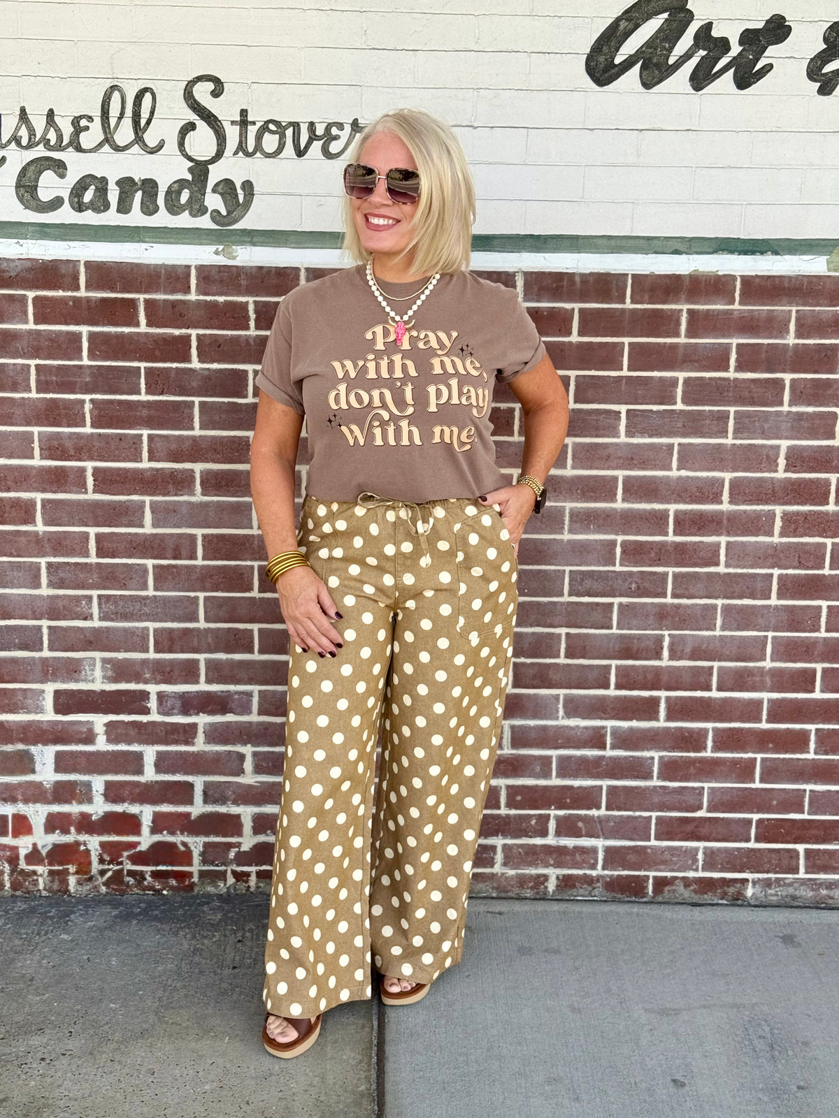 Unisex short sleeve brown shirt with 'Pray with Me Don't Play with Me' text, styled with patterned pants against a brick wall.