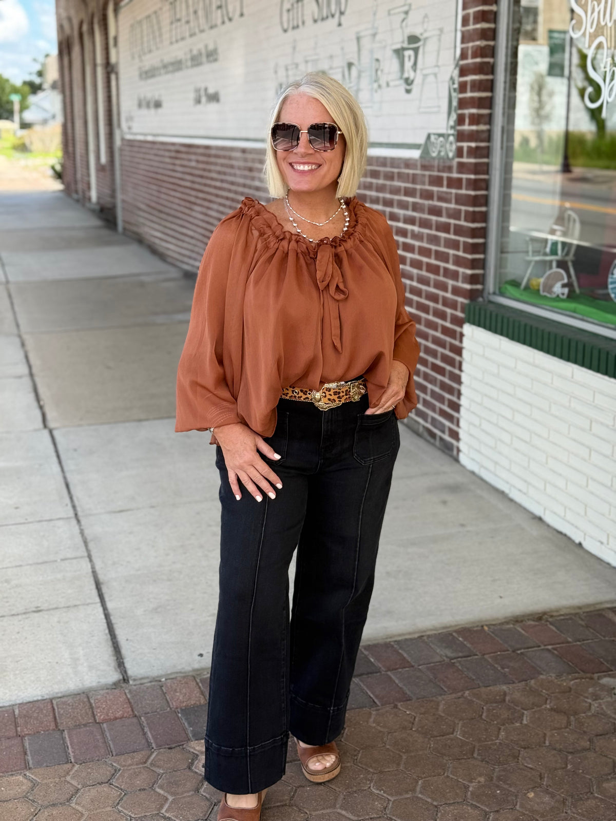 Rust-toned crinkle chiffon blouse with ruffled tie neck, styled with high-waisted black pants and accessories.