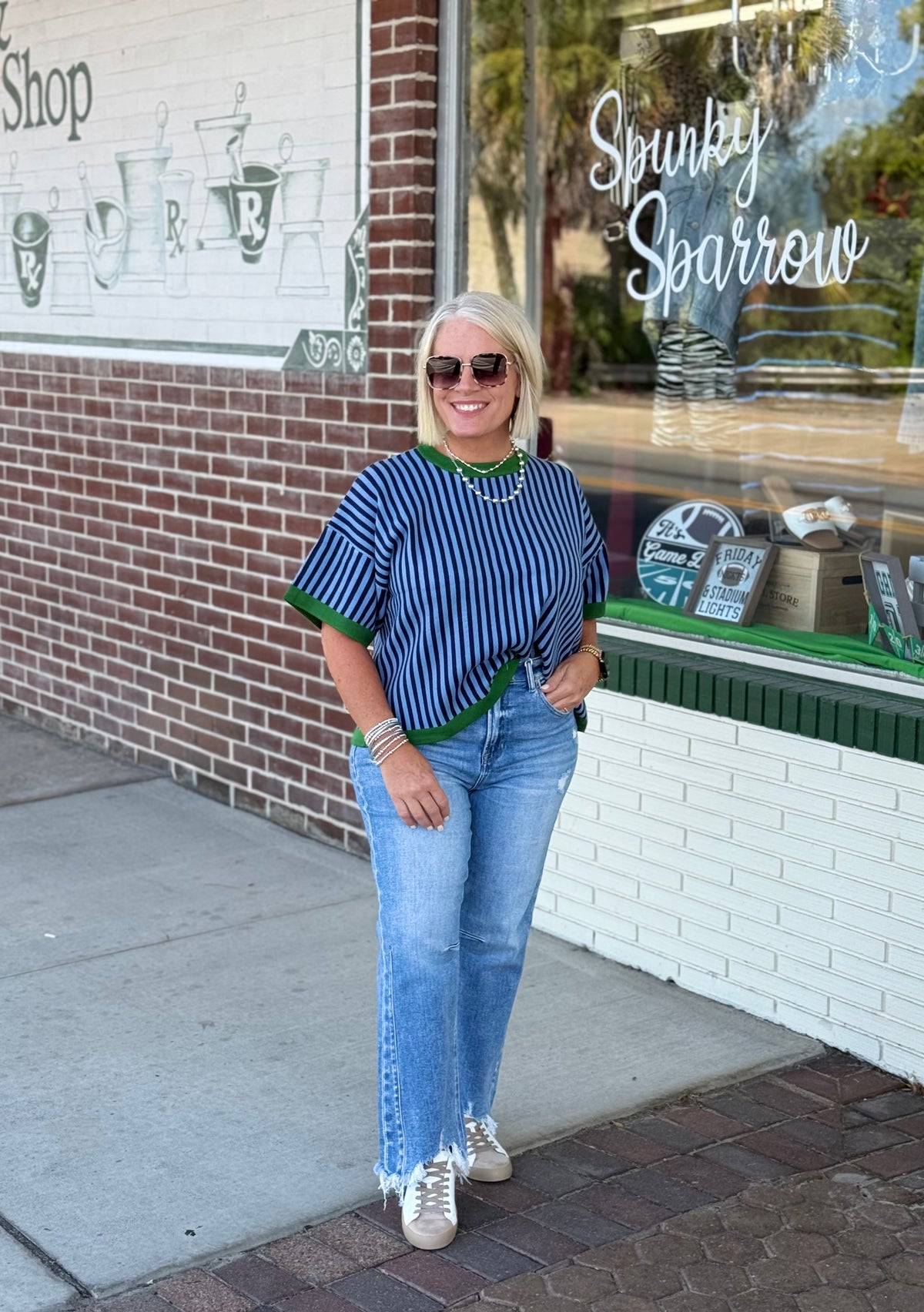 Model wearing a blue and green striped short sweater top with light blue jeans and casual sneakers against a brick wall.