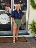 Olive green shorts with patch pockets and horsebit detail, styled with a navy top and tan sandals in a casual setting.