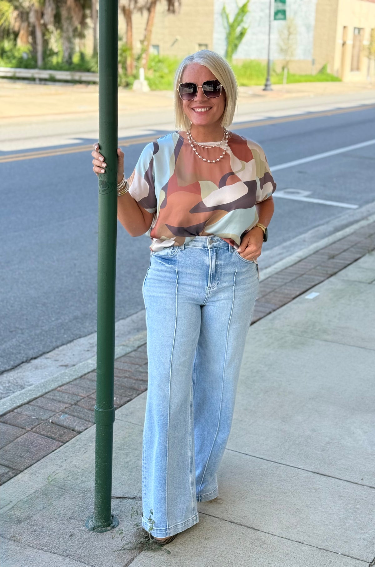 Model wearing a Multi Abstract Round Neck Short Sleeve Top with colorful design, paired with light blue wide-leg jeans outdoors.