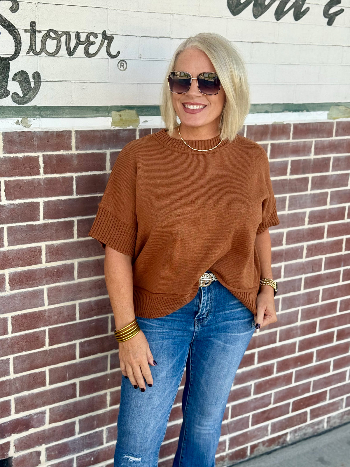 Drop sleeve sweater in caramel worn with blue jeans, featuring a crew neck and half sleeves against a brick wall backdrop.