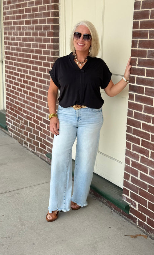 Black pleated blouse with short sleeves and collar, styled with light blue wide-leg jeans and brown sandals against a brick wall.