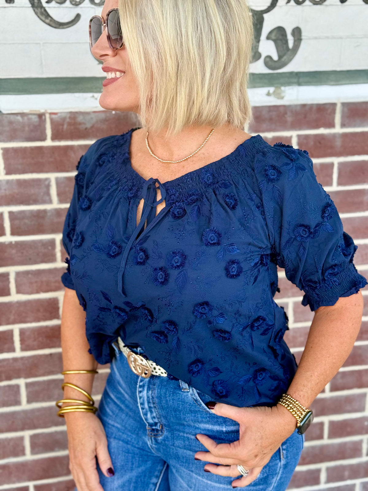 Navy blouse with 3D floral embroidery, boatneck neckline, and gathered puff sleeves styled with high-waisted jeans.