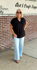 Black pleated blouse with collar and short sleeves, styled with light blue flared jeans and accessories against a brick wall.