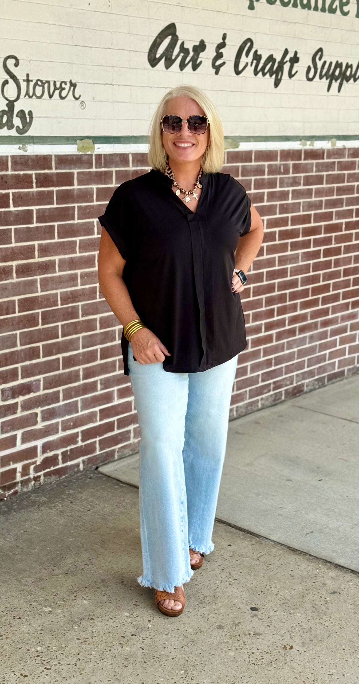 Black pleated blouse with collar and short sleeves, styled with light blue flared jeans and accessories against a brick wall.