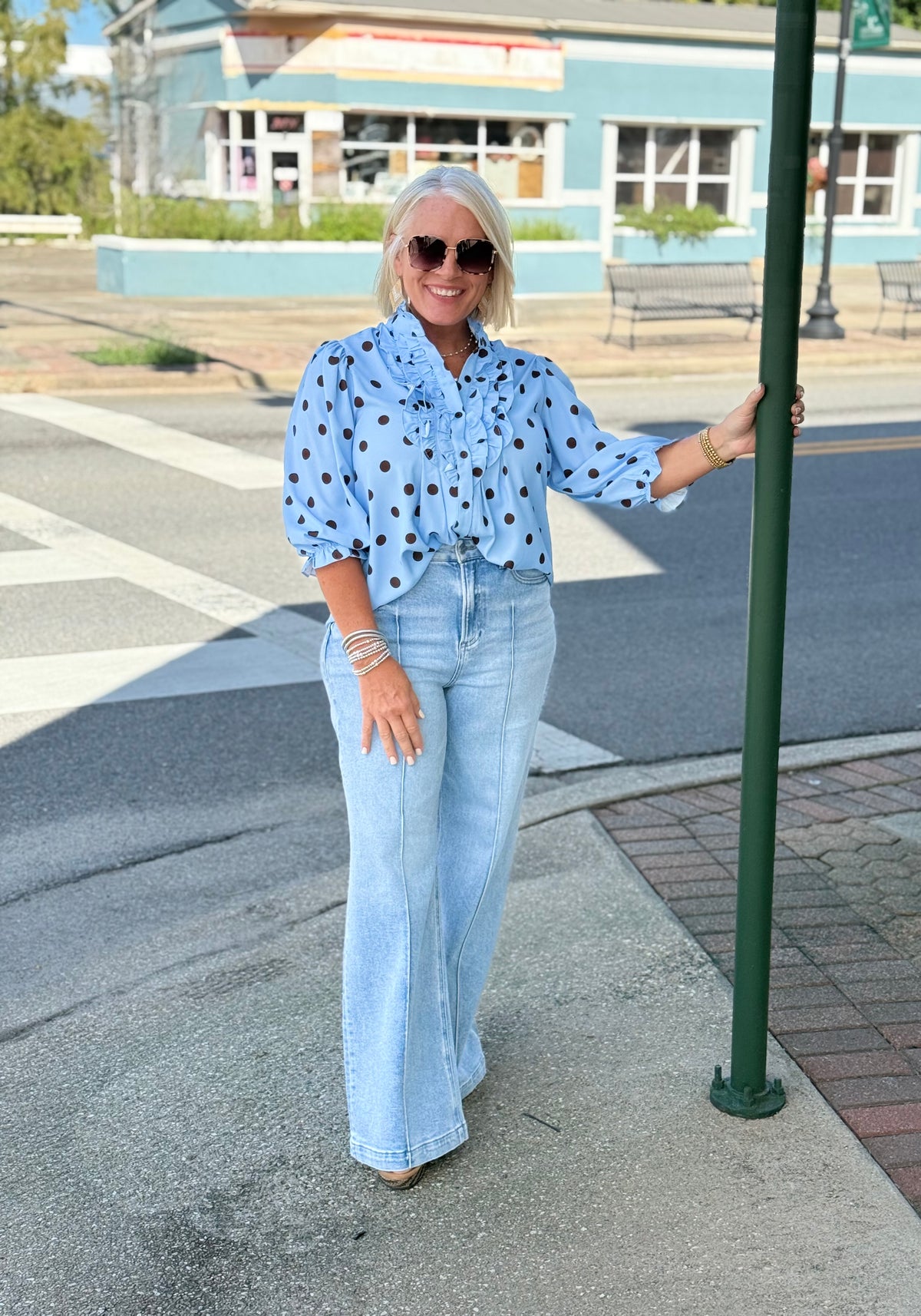 Blue polka dot ruffle blouse with long sleeves, styled casually with light blue flared jeans and sunglasses outdoors.