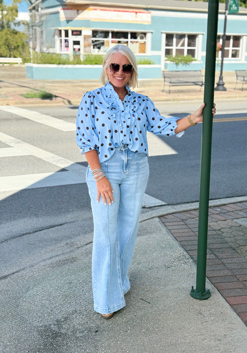 Blue polka dot ruffle blouse with long sleeves, styled casually with light blue flared jeans and sunglasses outdoors.
