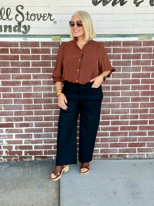Person wearing black cropped wide-leg pants with scallop pockets, styled with a brown top against a brick wall background.