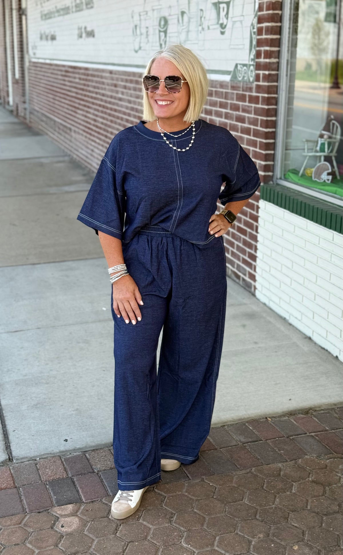 Model wearing a denim blue half-sleeve top with top stitch detailing and matching loose-fitting long pants, accessorized with sunglasses.