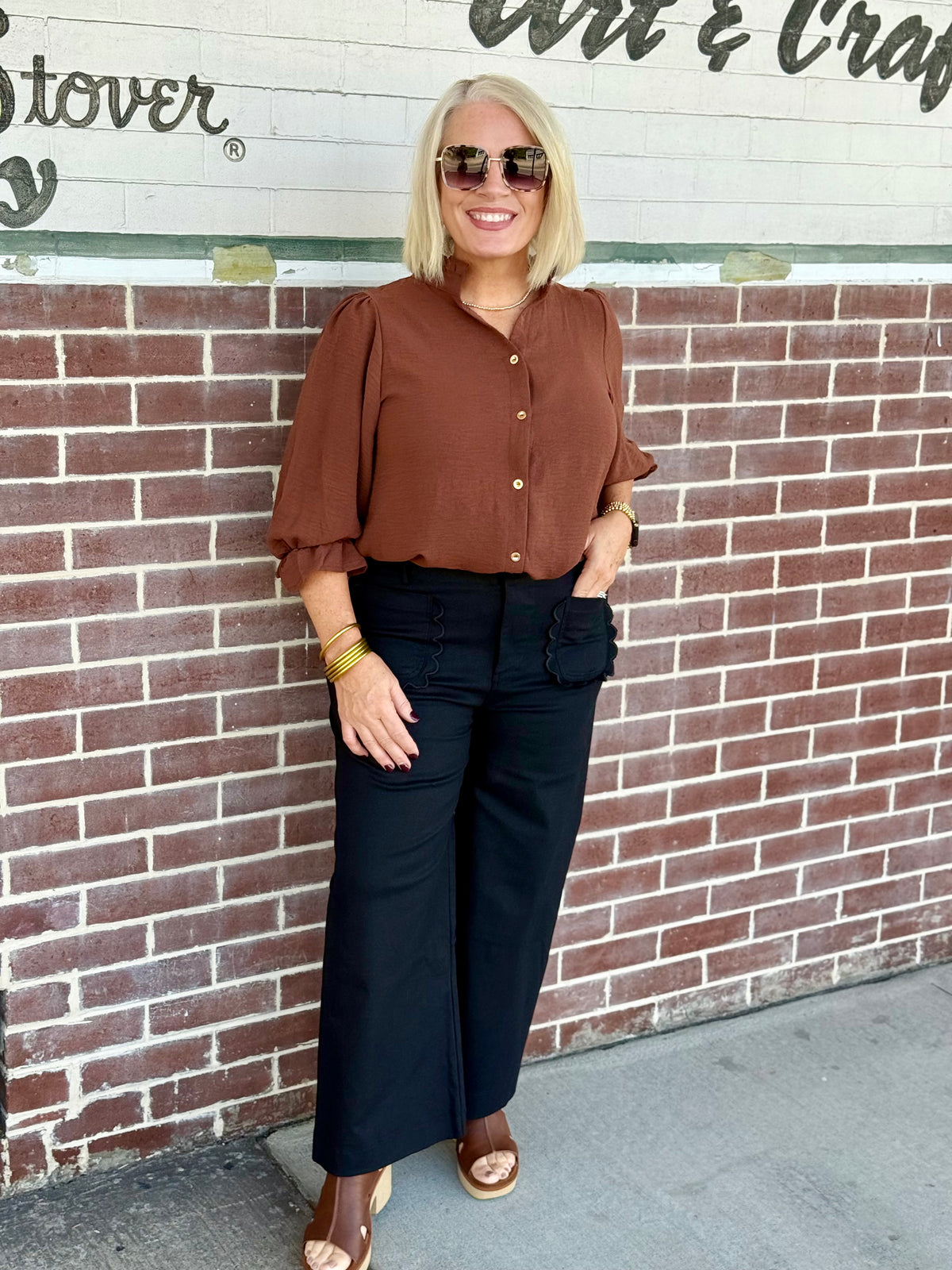 Person wearing black cropped wide-leg pants with scallop pockets, styled with a brown top against a brick wall.