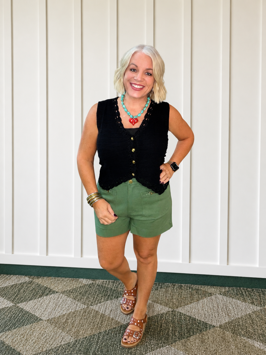 Black crochet trimmed button-down sweater vest styled over green bottoms with a colorful necklace against a neutral background.