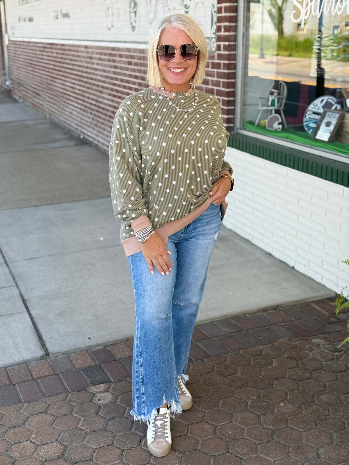 Olive top with polka dot print, relaxed drop shoulder silhouette, and triangle cutout detail, styled with light blue jeans.