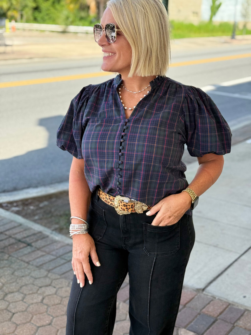 Navy plaid blouse with V-neckline and button-down front, featuring soft puff sleeves, styled with a belt and black pants.