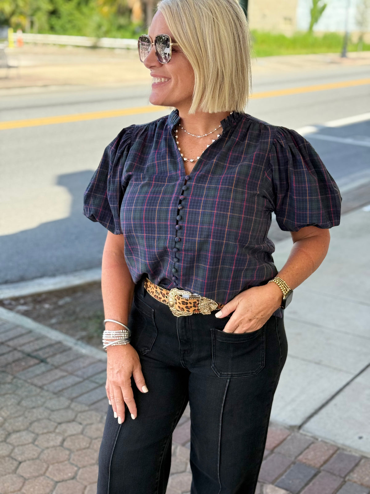 Navy plaid blouse with V-neckline and button-down front, featuring soft puff sleeves, styled with a belt and black pants.
