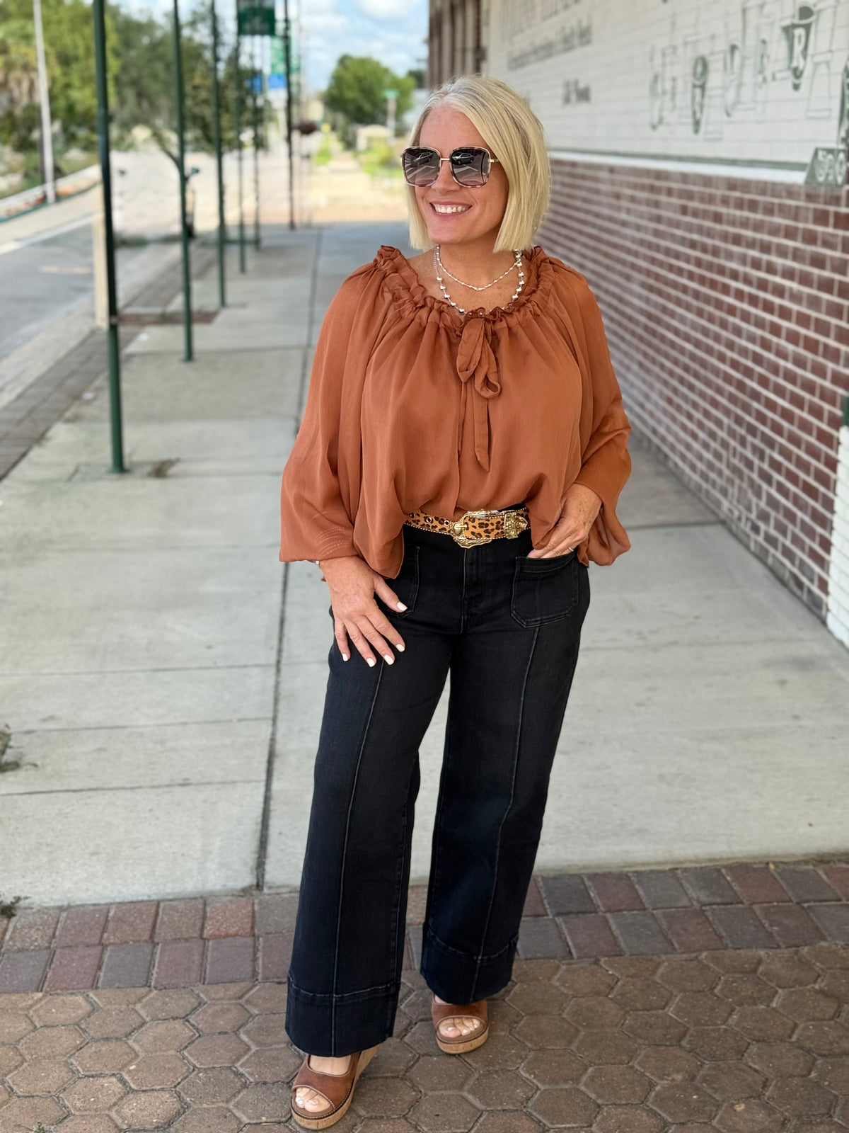 Rust-toned blouse with ruffled tie neck, styled with high-waisted black pants and sunglasses outdoors.