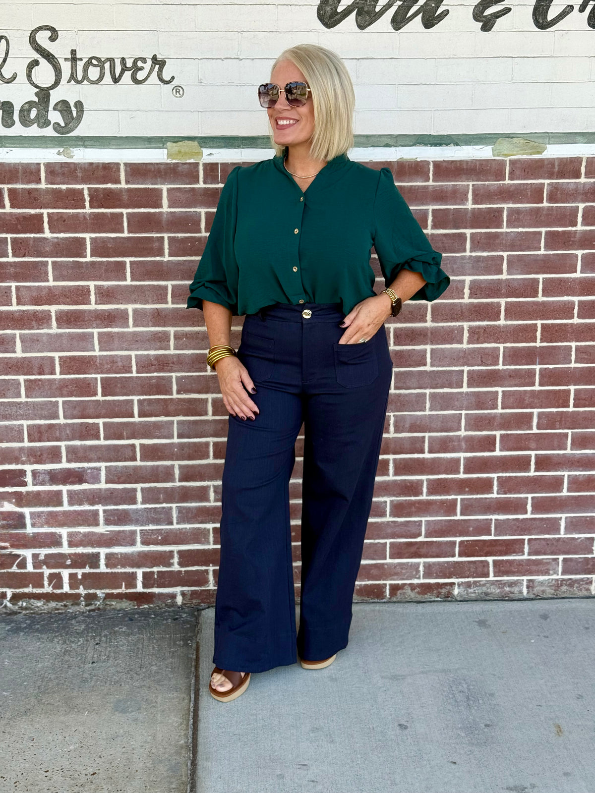 Model wearing a hunter green 3/4 sleeve button-down blouse styled with high-waisted wide-leg pants against a brick wall.