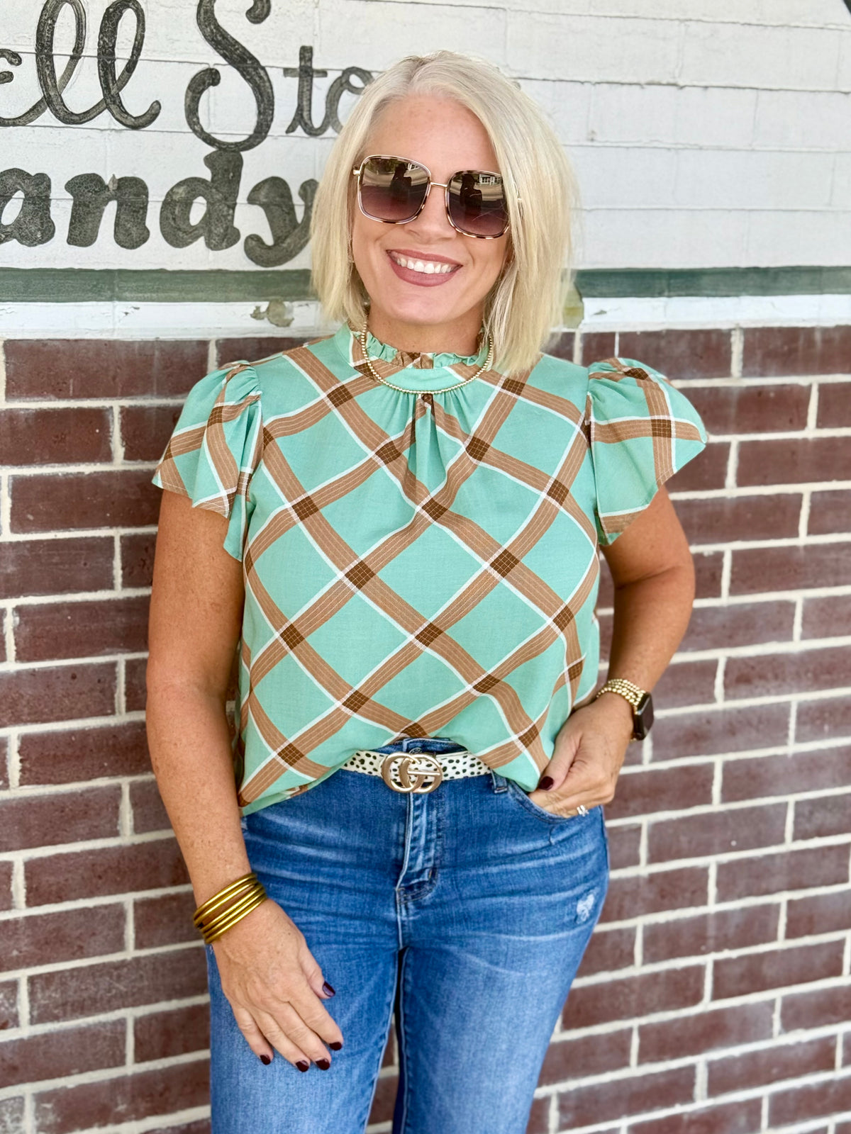 Sage plaid blouse with ruffle sleeves and round neckline, styled with blue jeans and a belt.