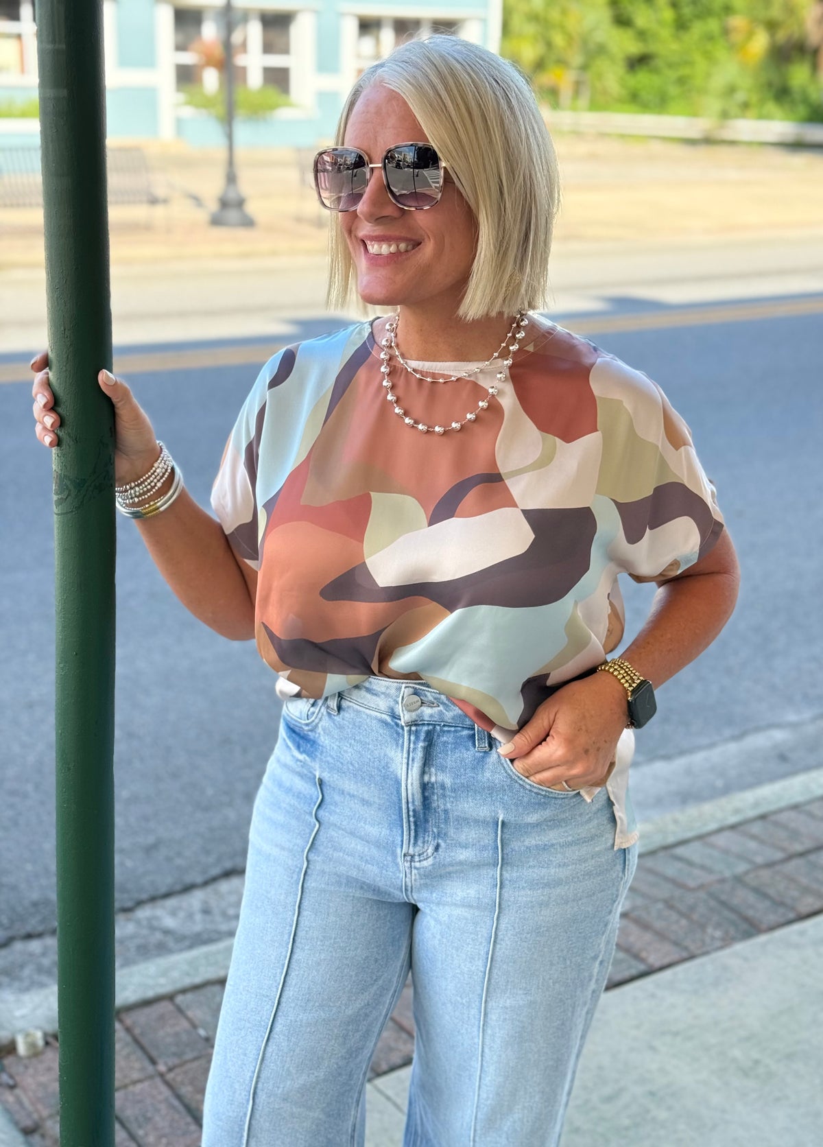 Model wearing a multi-colored abstract round neck top with short sleeves, paired with light blue high-waisted jeans and accessories.