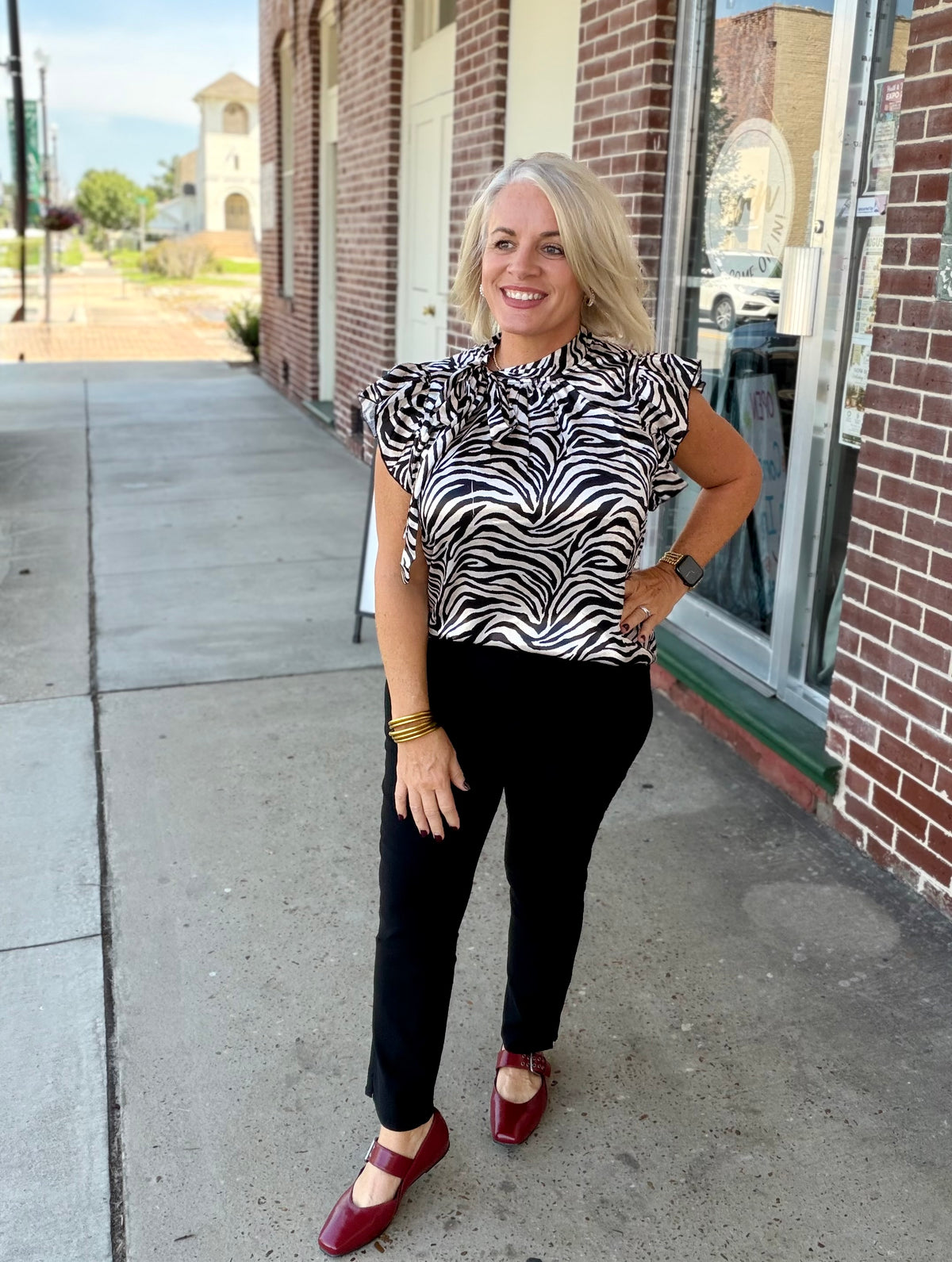 Bold zebra print top with functional tie neckline, short ruffled sleeves, styled with black pants and red shoes outdoors.