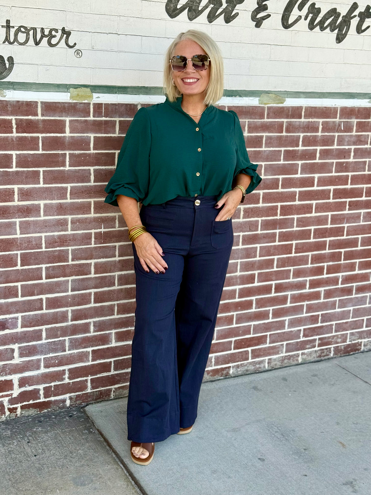 Model wearing a hunter green 3/4 sleeve button-down blouse styled with high-waisted wide-leg pants against a brick wall.
