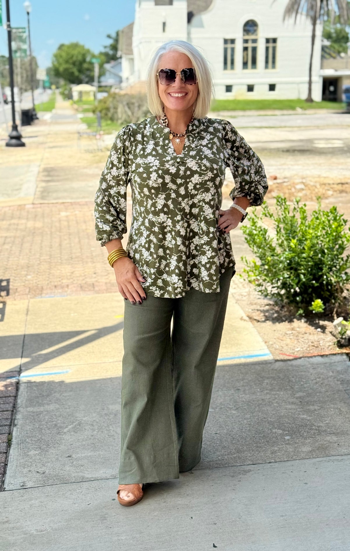 Dark olive blouse with floral pattern and smocked sleeves, styled with wide-leg trousers outdoors.