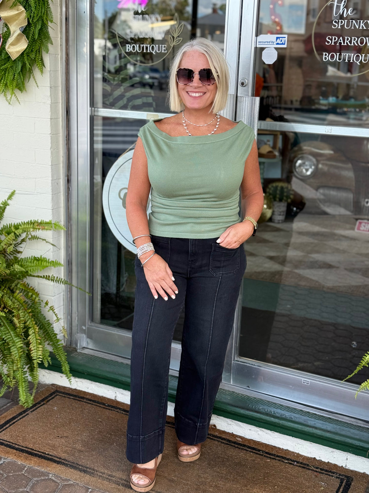 Sage wide neck pleated knit top styled with black trousers, worn by a model outside a boutique.