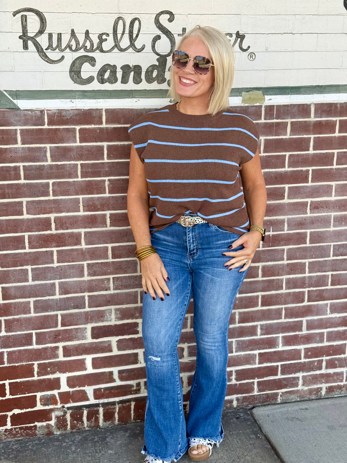 Chocolate brown sweater vest with blue stripes, relaxed fit, short sleeves, styled with flared jeans against a brick wall.
