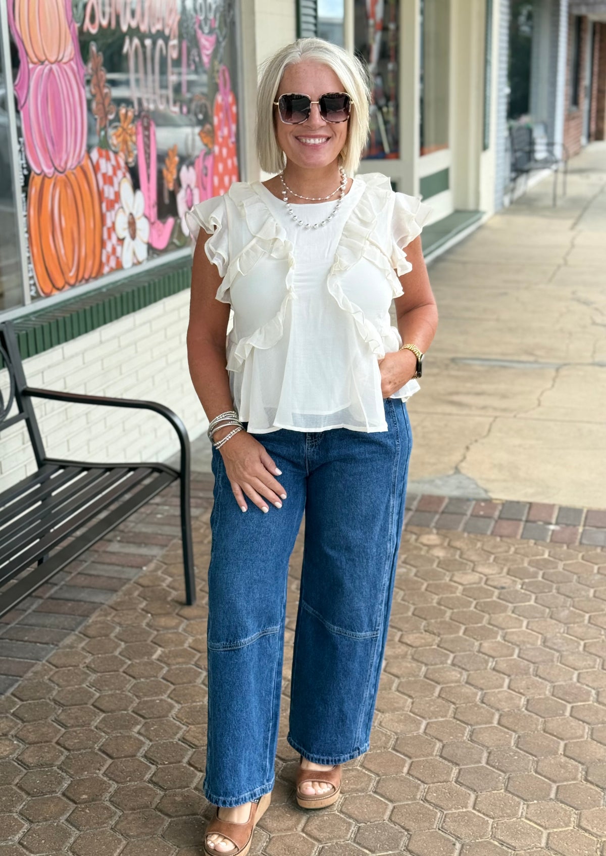 Cream ruffled blouse styled with high-waisted wide-leg jeans, model wearing sunglasses and jewelry outdoors.