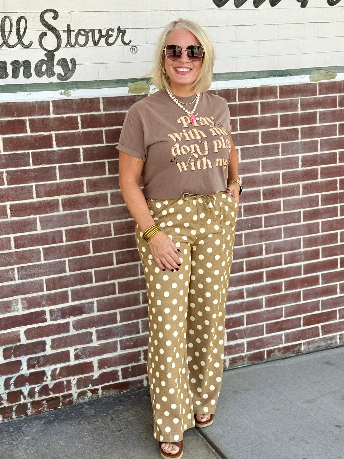 Taupe relaxed wide-leg polka dot drawstring pants styled casually with a brown graphic t-shirt against a brick wall.