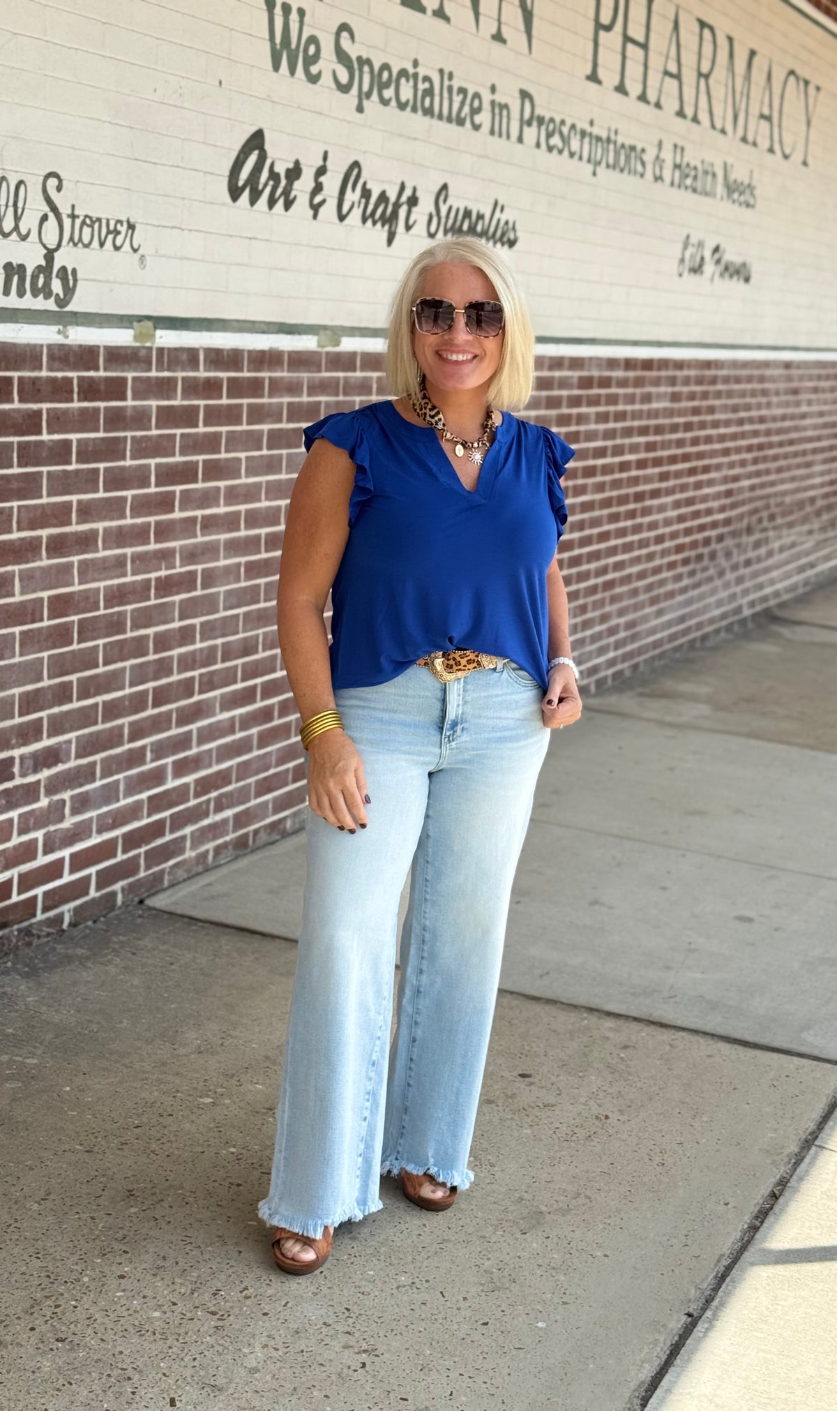 Model wearing a royal blue flutter sleeve top with a v-neckline, paired with light blue flared jeans and brown sandals outdoors.