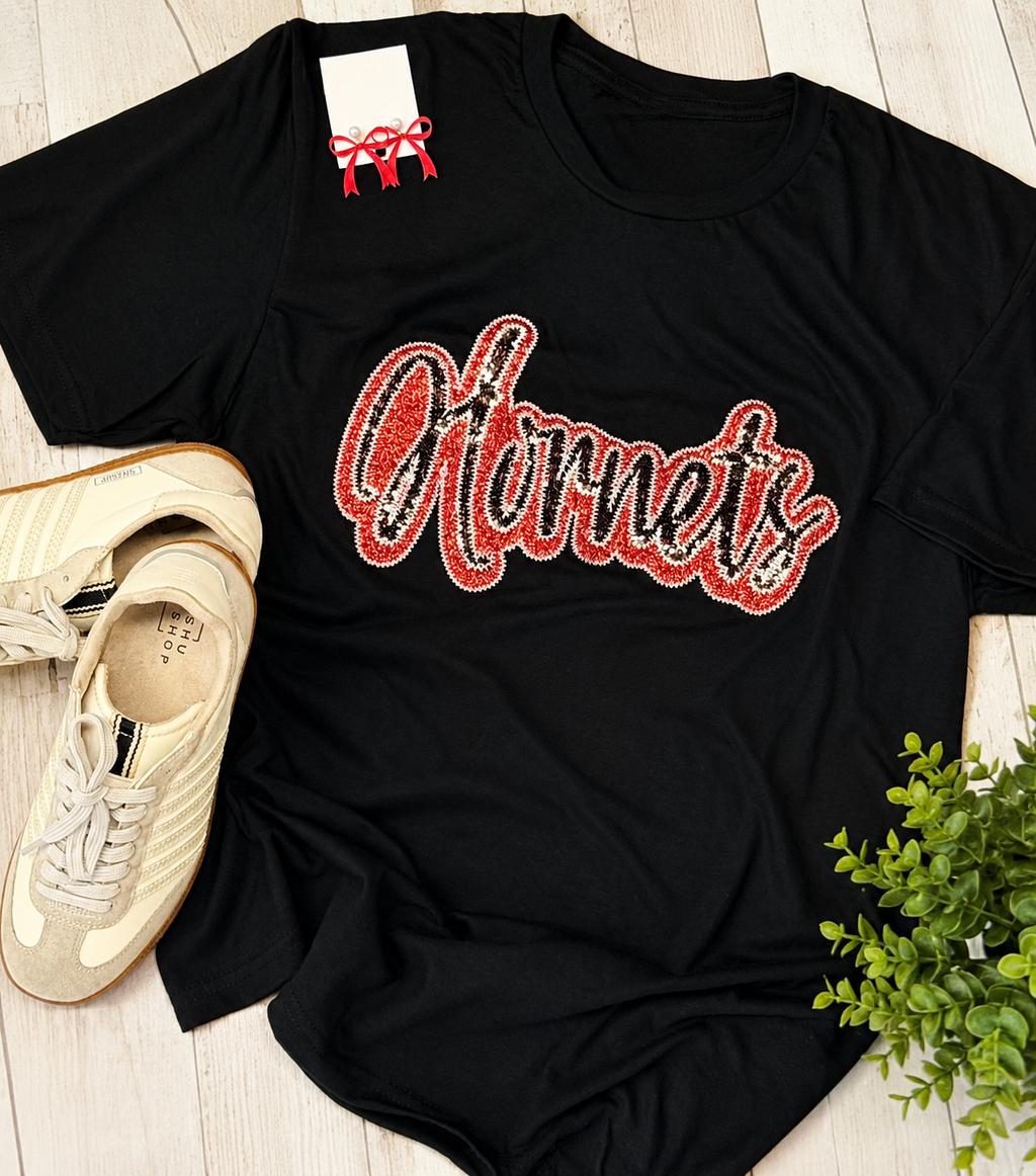 Black short sleeve t-shirt with sequined 'Hornets' text, styled with sneakers and decorative items on wooden flooring.