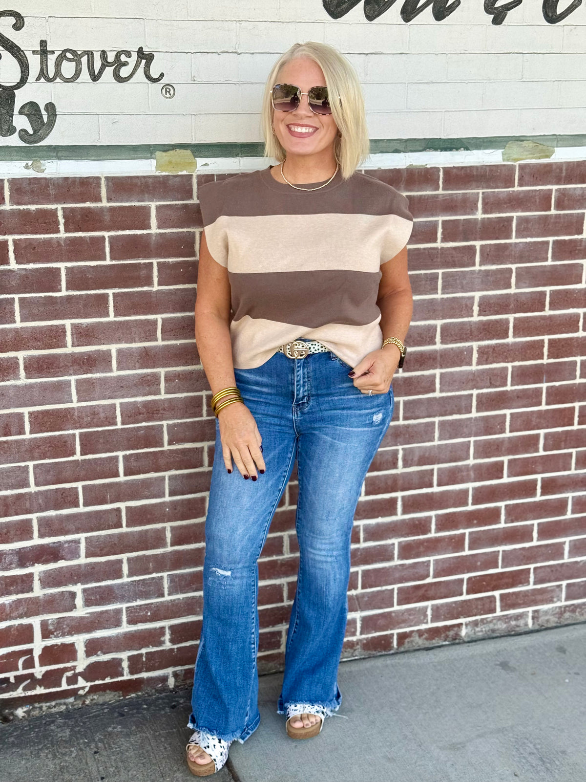 Sleeveless mocha and cream striped sweater top styled with blue jeans and accessories against a brick wall.