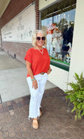 Model wearing a vibrant orange short sleeve top with a relaxed fit, paired with white pants in an outdoor setting.