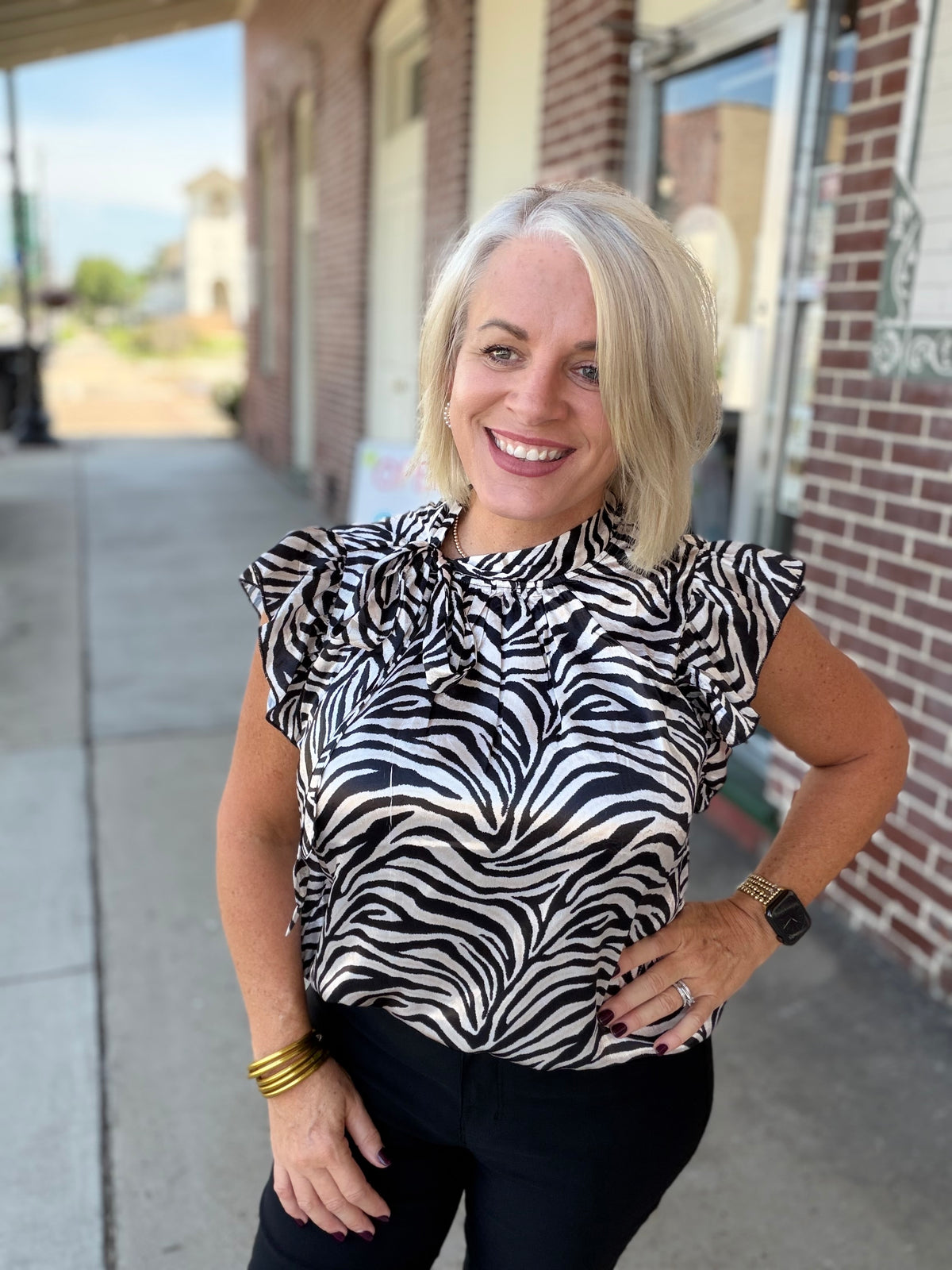 Model wearing the Savanna Stripe Top with bold zebra print and a functional tie neckline, styled with black pants outdoors.