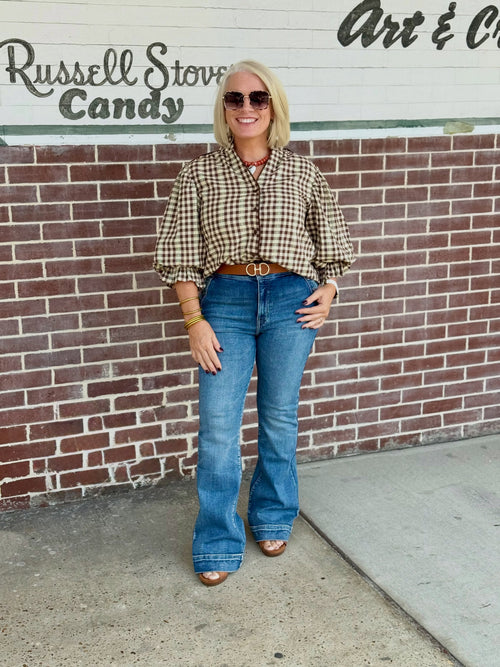 Brown check print button-down shirt with three-quarter gathered balloon sleeves styled with a belt and flared jeans against a brick wall.